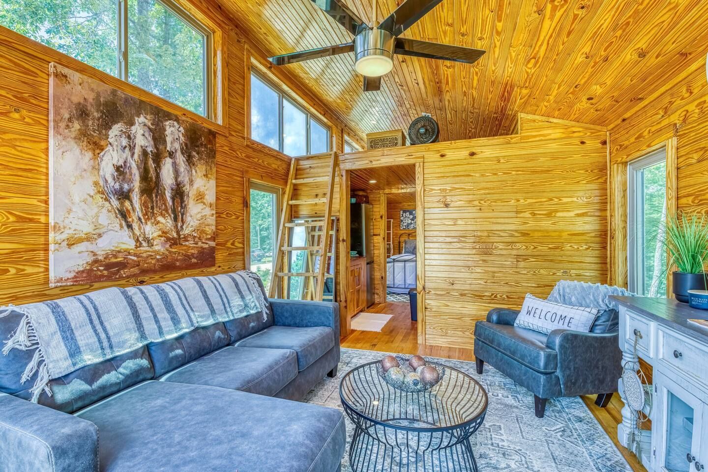 Cozy cabin interior: wood paneling, couch, armchair, art, small table, ladder to loft.