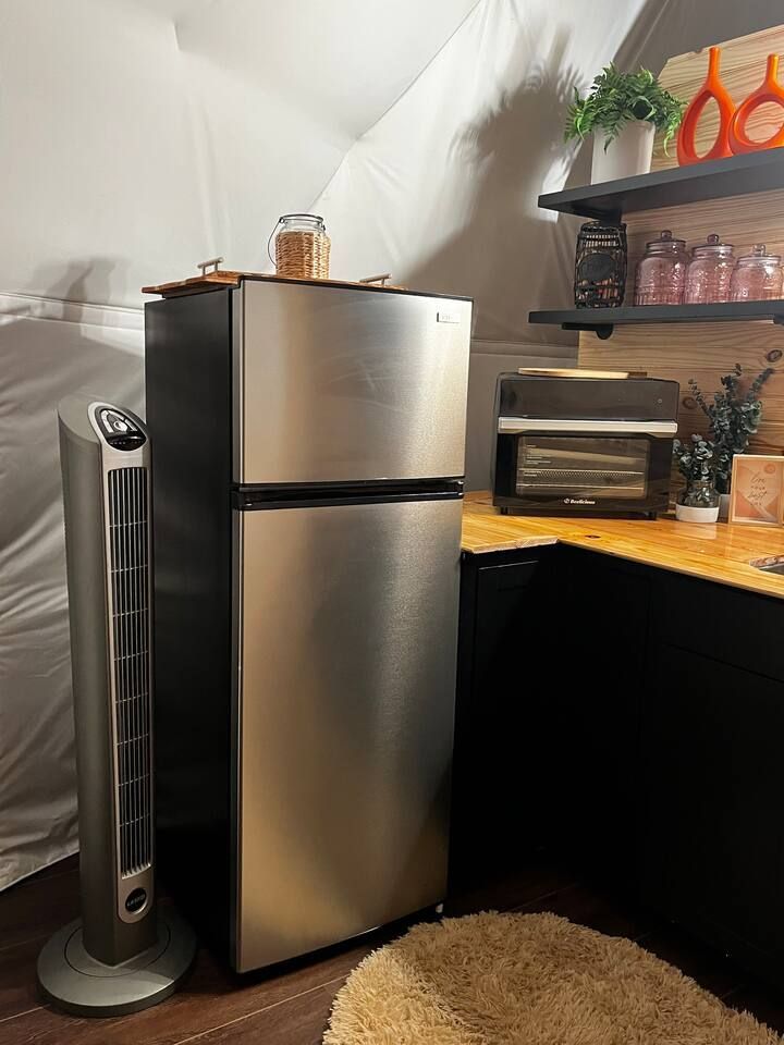 Stainless steel refrigerator in a room with a fan, counter, and shelves with jars and decor.