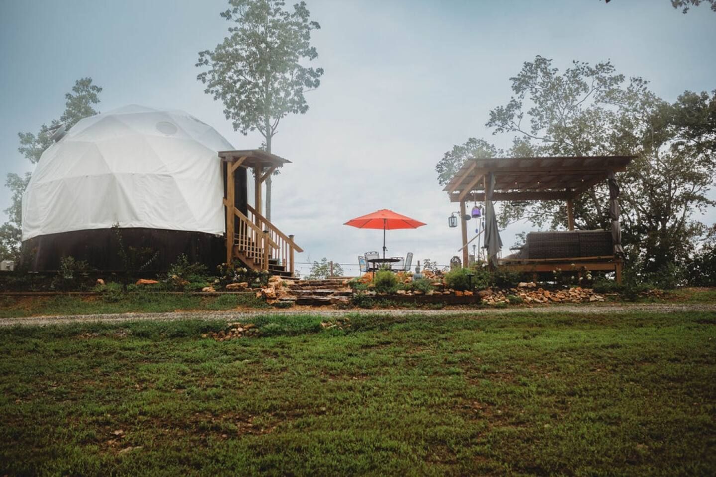 Geodesic dome structure with a deck, patio, and orange umbrella in a green landscape under a cloudy sky.