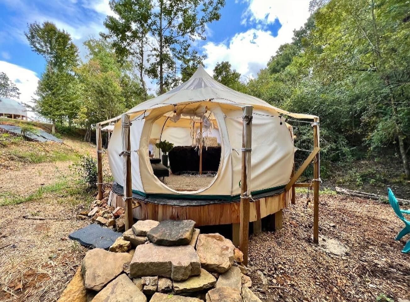 A canvas glamping tent with wooden supports, on a wooden platform, surrounded by trees and a rocky path.