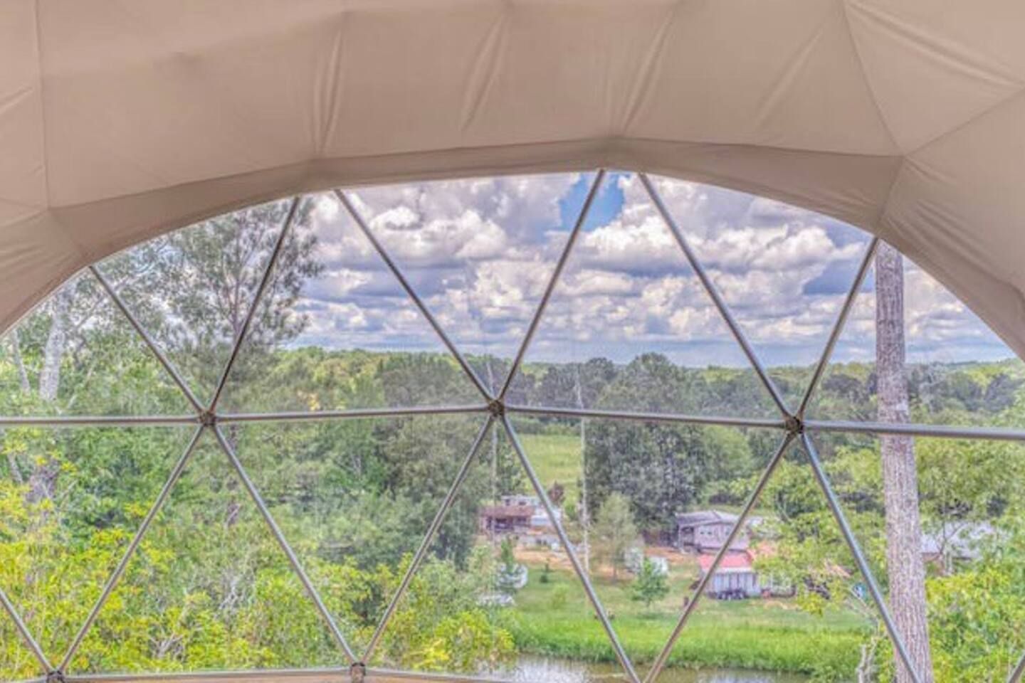 View from a geodesic dome window looking at trees, a small pond, and houses under a cloudy sky.