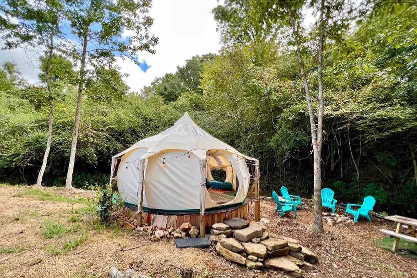 A glamping tent in a forest setting with Adirondack chairs and a picnic table.