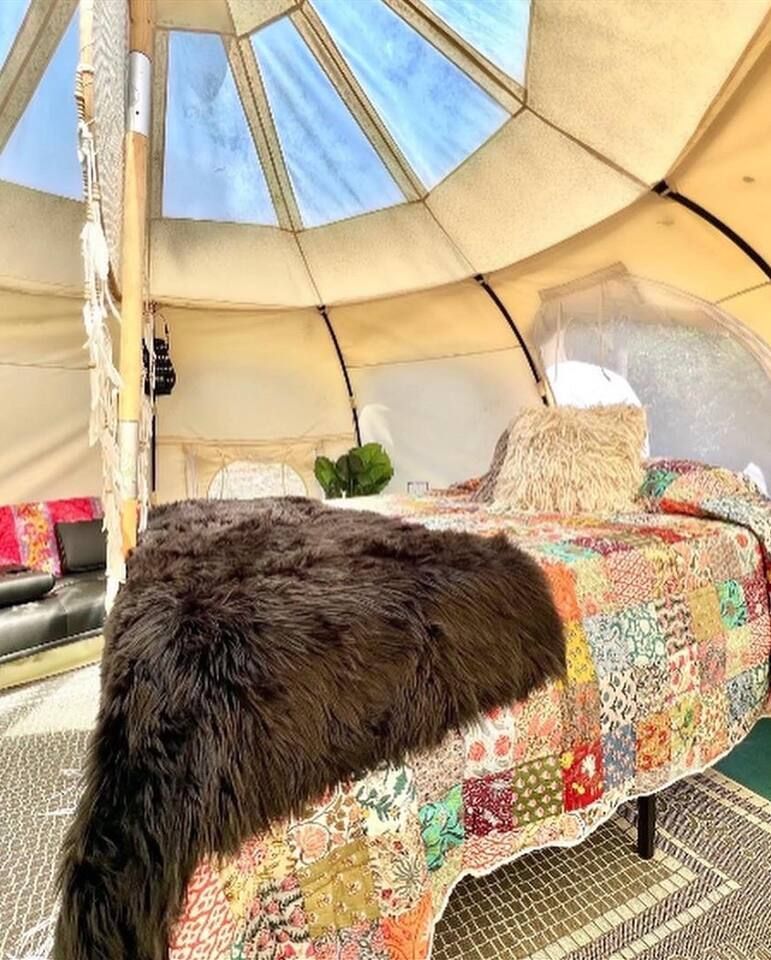 Inside a beige teepee: a bed with a patchwork quilt, brown fur throw, and small pillow. Skylight overhead.