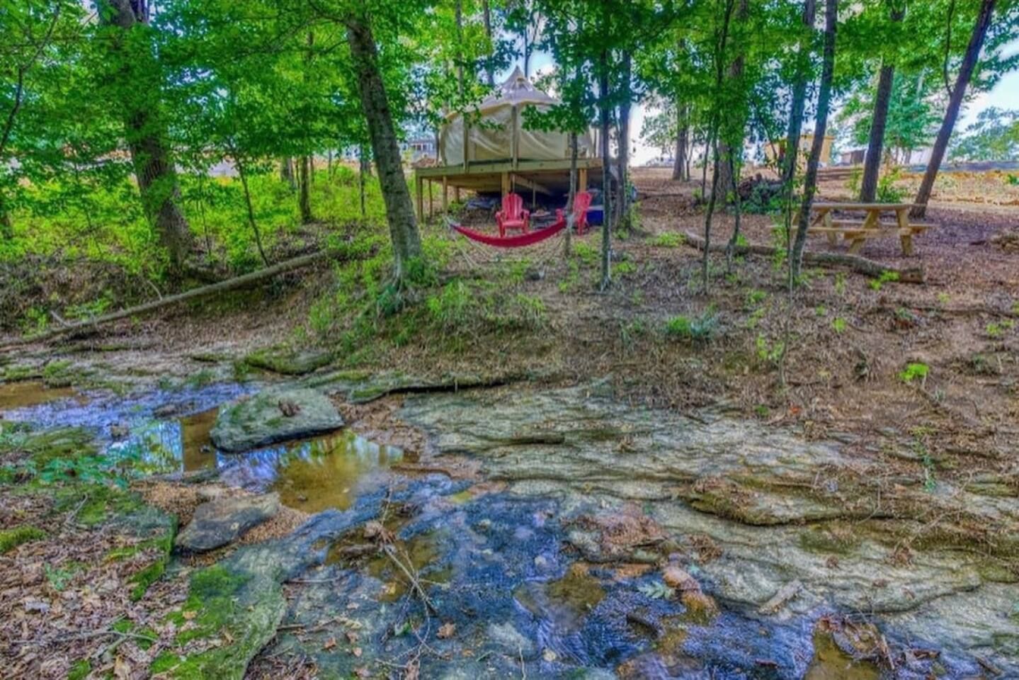 Creek bed in foreground leads to a deck with a red hammock and treehouse in a wooded setting.