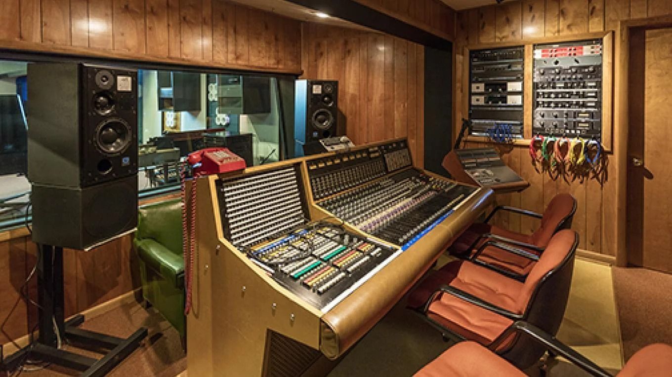 Recording studio control room with mixing console, monitors, and sound booth visible through window.