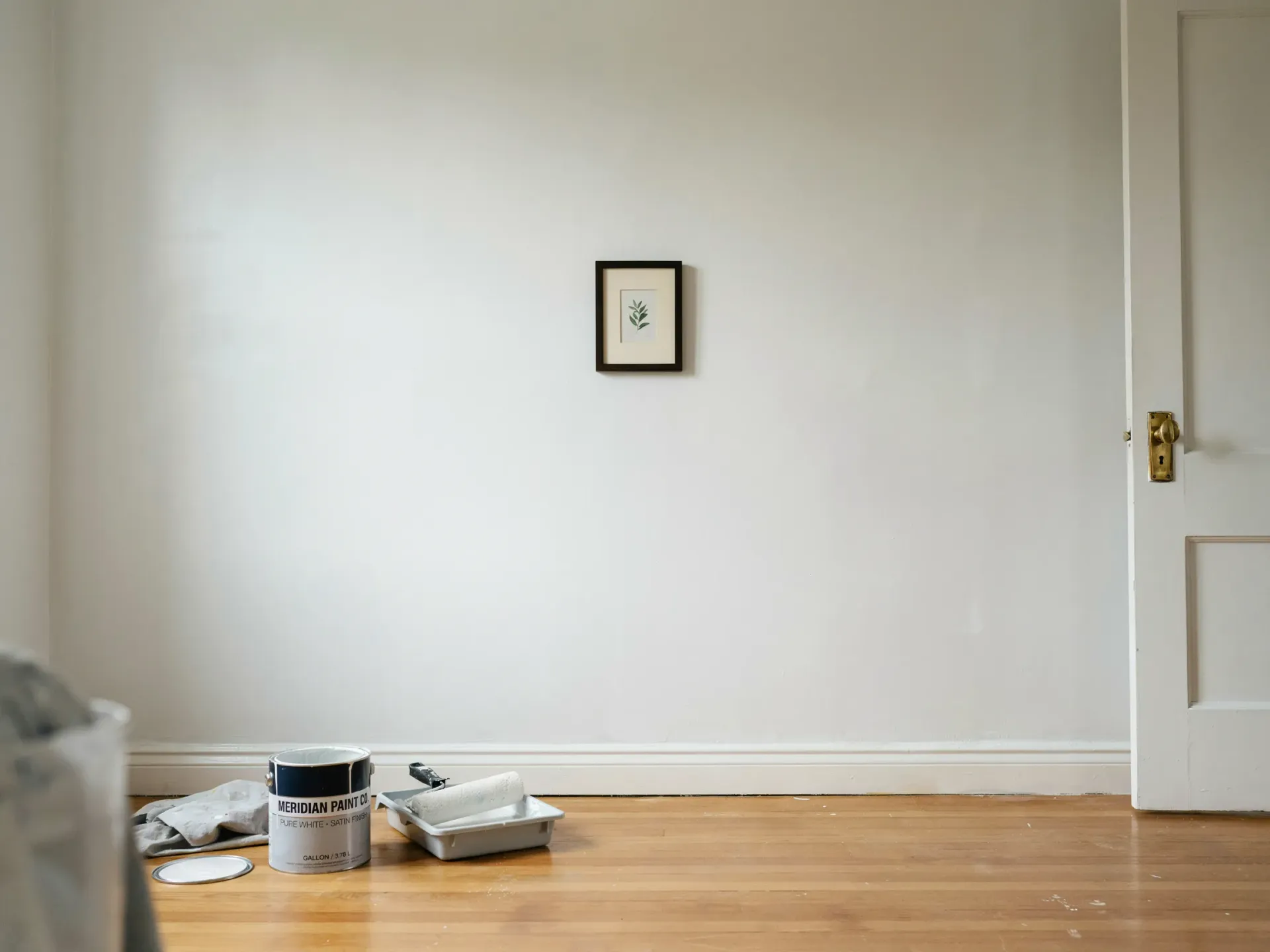 Empty white room with a framed picture on the wall, paint supplies on the floor, and a partially open door