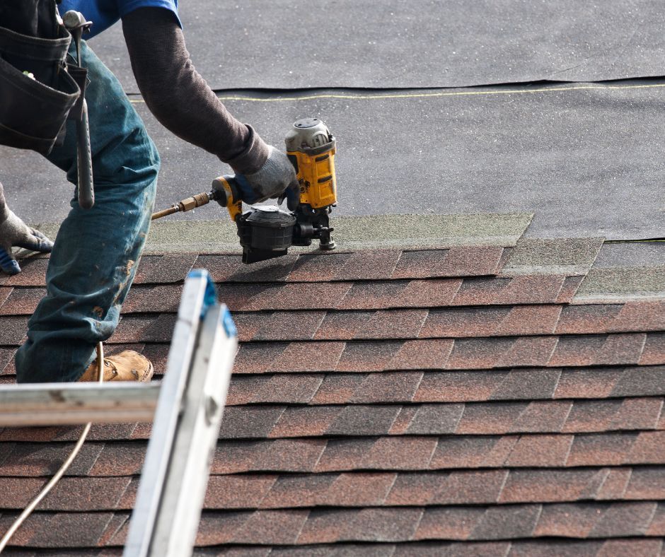 Worker using a nail gun to repair a shingled roof beside a ladder