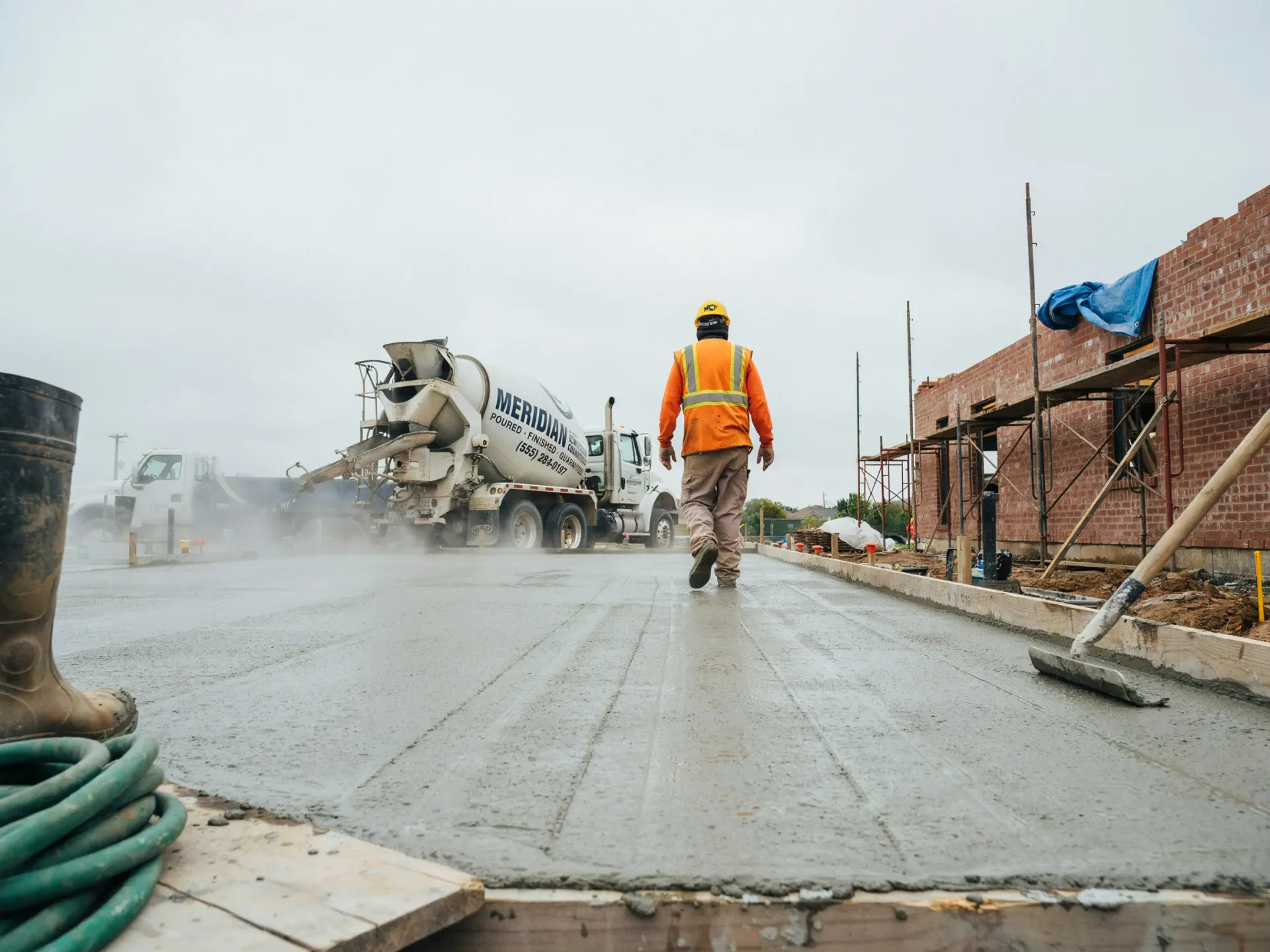 Construction worker walks on wet road near cement mixer at roadside worksite.