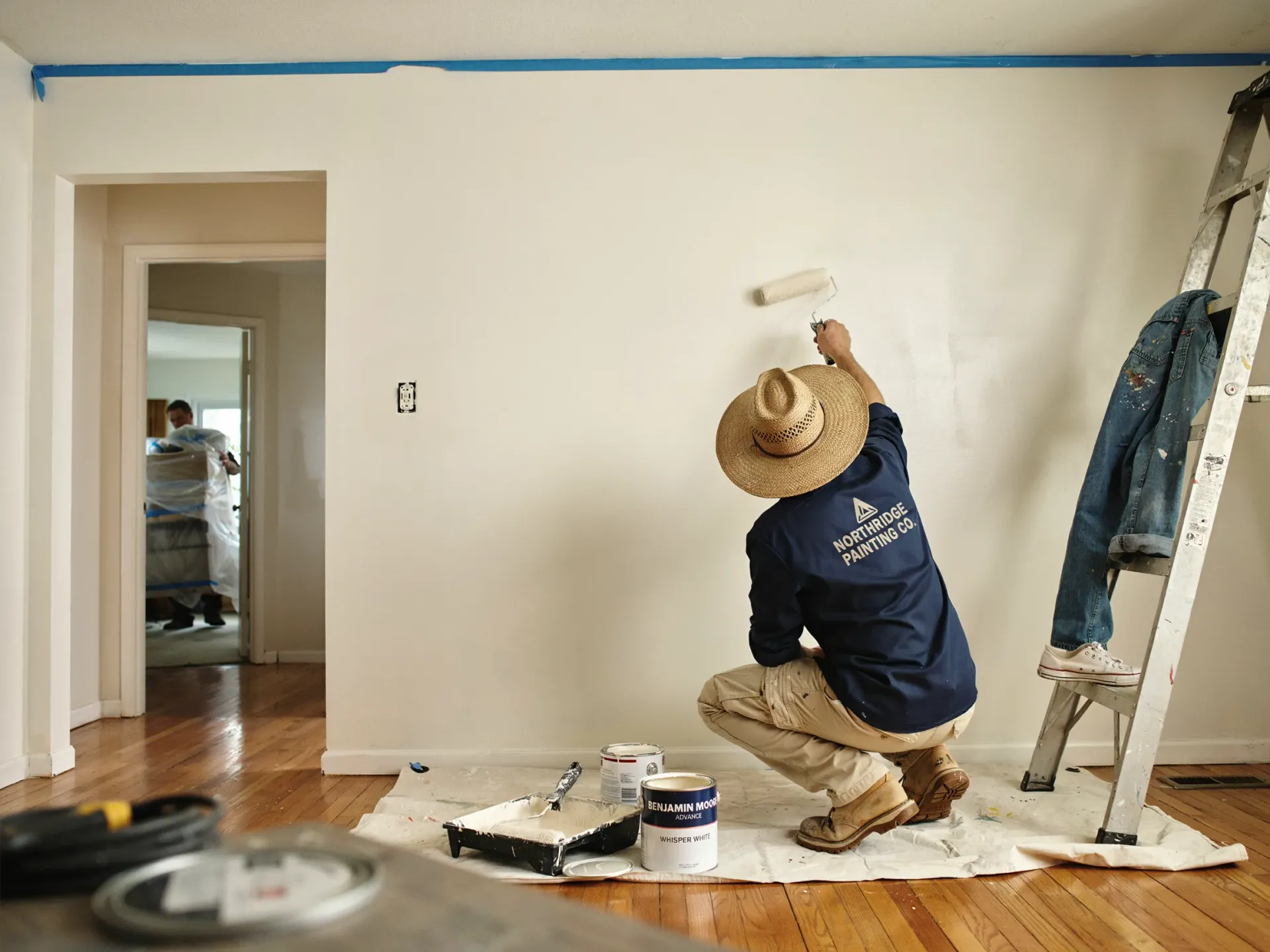 Painter in blue shirt and straw hat painting a white wall in a room with drop cloths and a ladder.