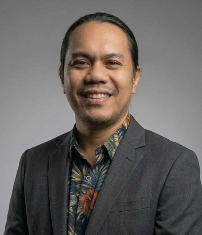 Man with dark hair pulled back, wearing a blue blazer and gray sweater, looking directly at the camera.
