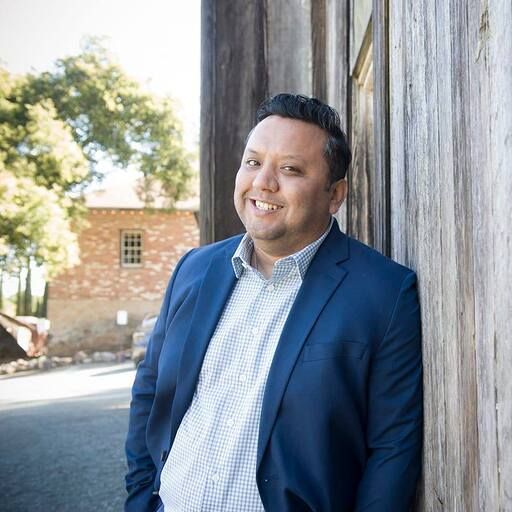 Man in a blue blazer smiles, leaning against a weathered wooden wall, with a building and tree in the background.