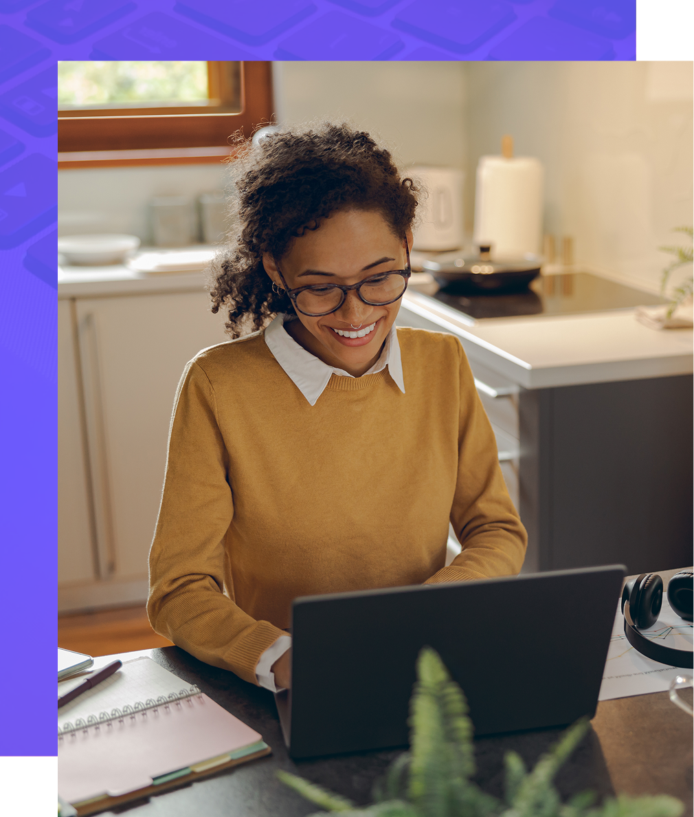 Woman with glasses, yellow sweater, working on a laptop at a kitchen table, smiling.