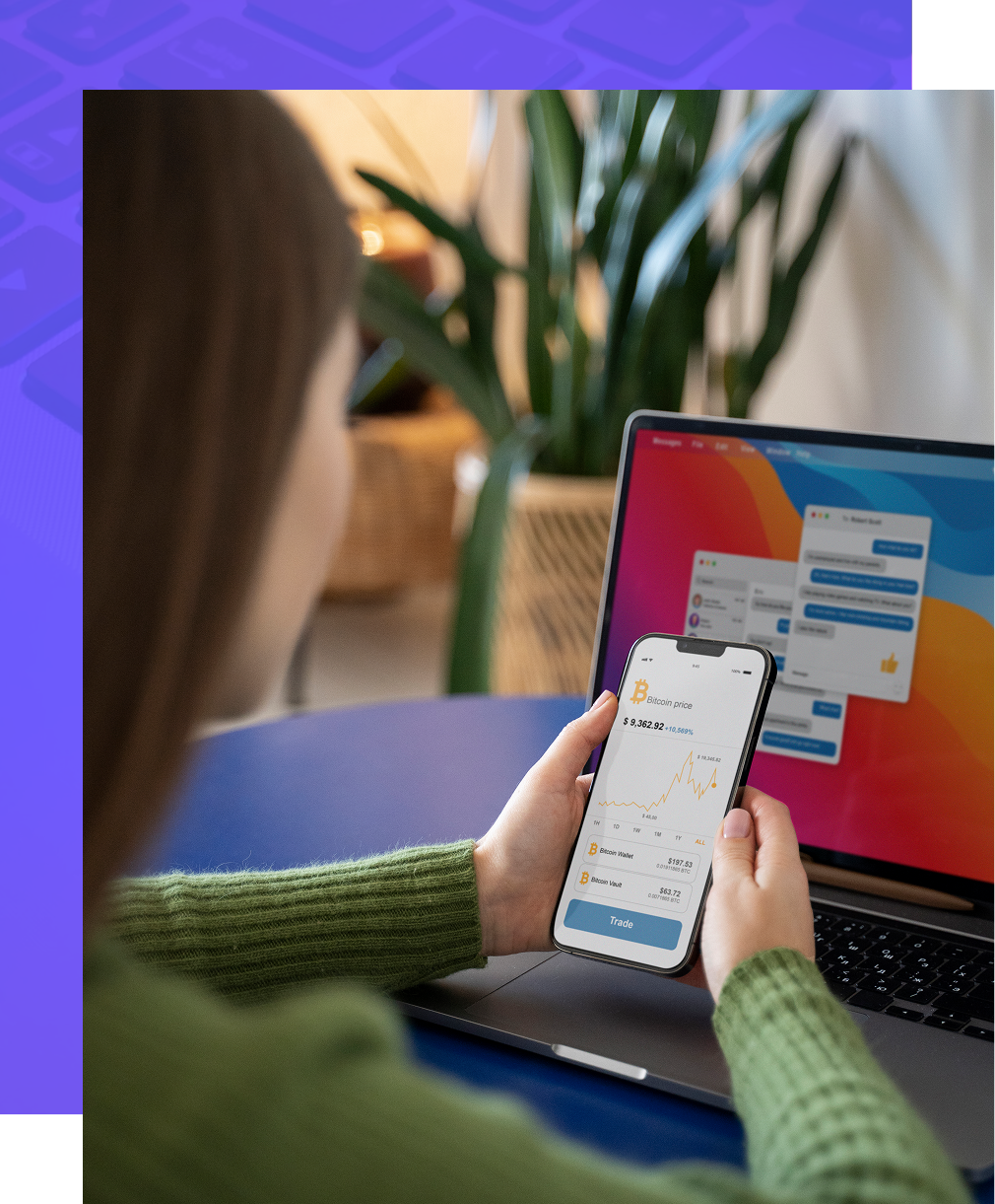 Person in a green sweater holding a smartphone with an app interface while working on a laptop at a table.