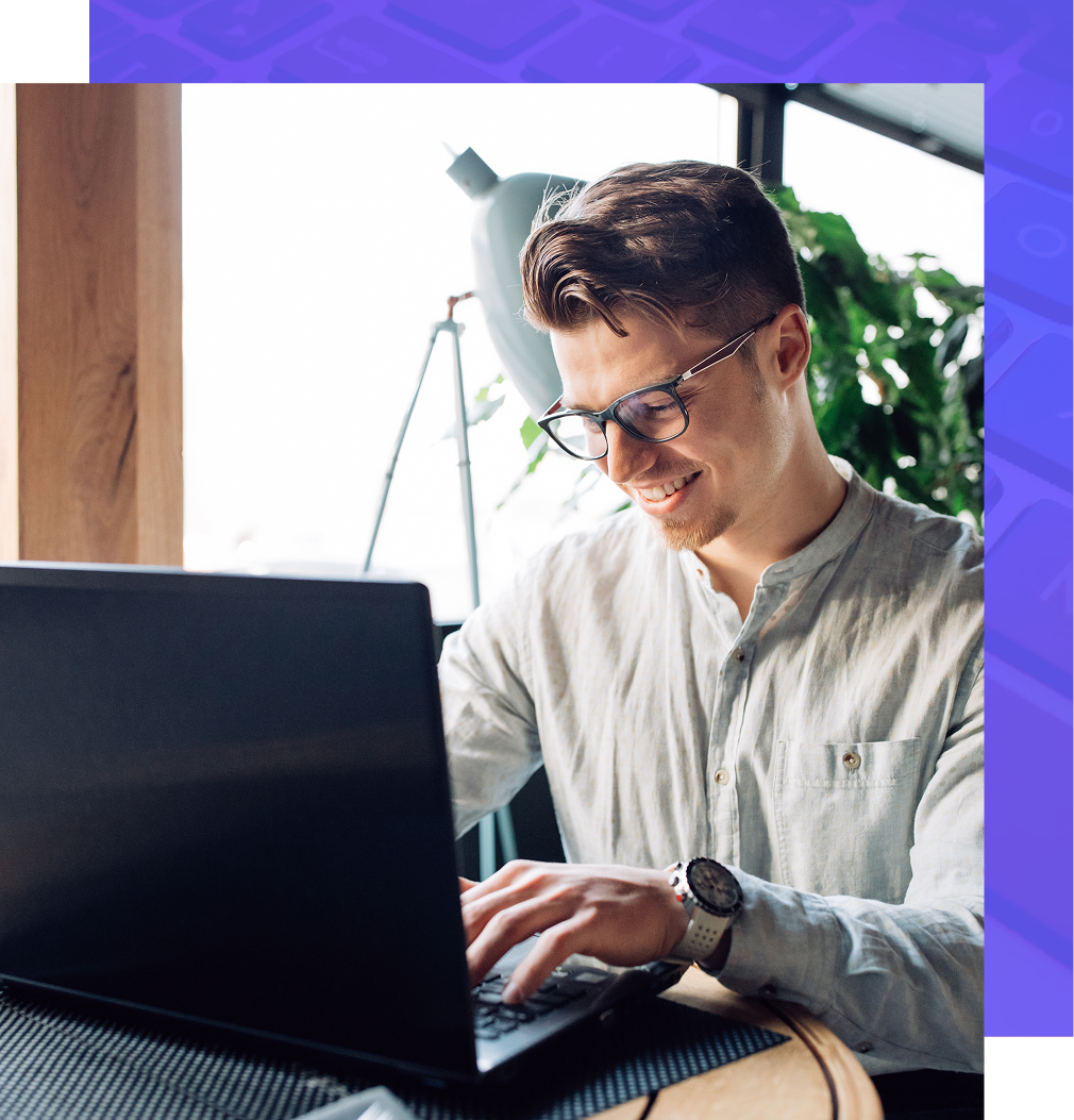 A person wearing glasses and a button-up shirt smiling while typing on a laptop in a bright, plant-filled workspace.