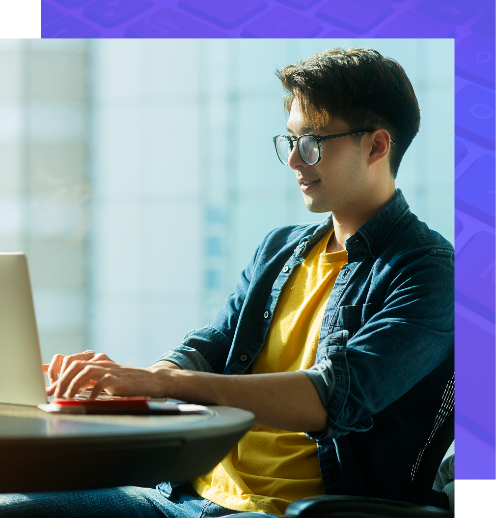 A person wearing glasses and a yellow shirt over a denim shirt sits at a desk typing on a laptop in a bright office.