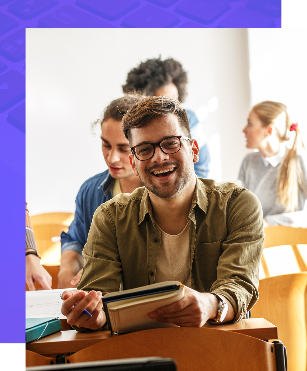 A smiling student in an olive green shirt holds a notebook while sitting at a desk in a classroom with other students.
