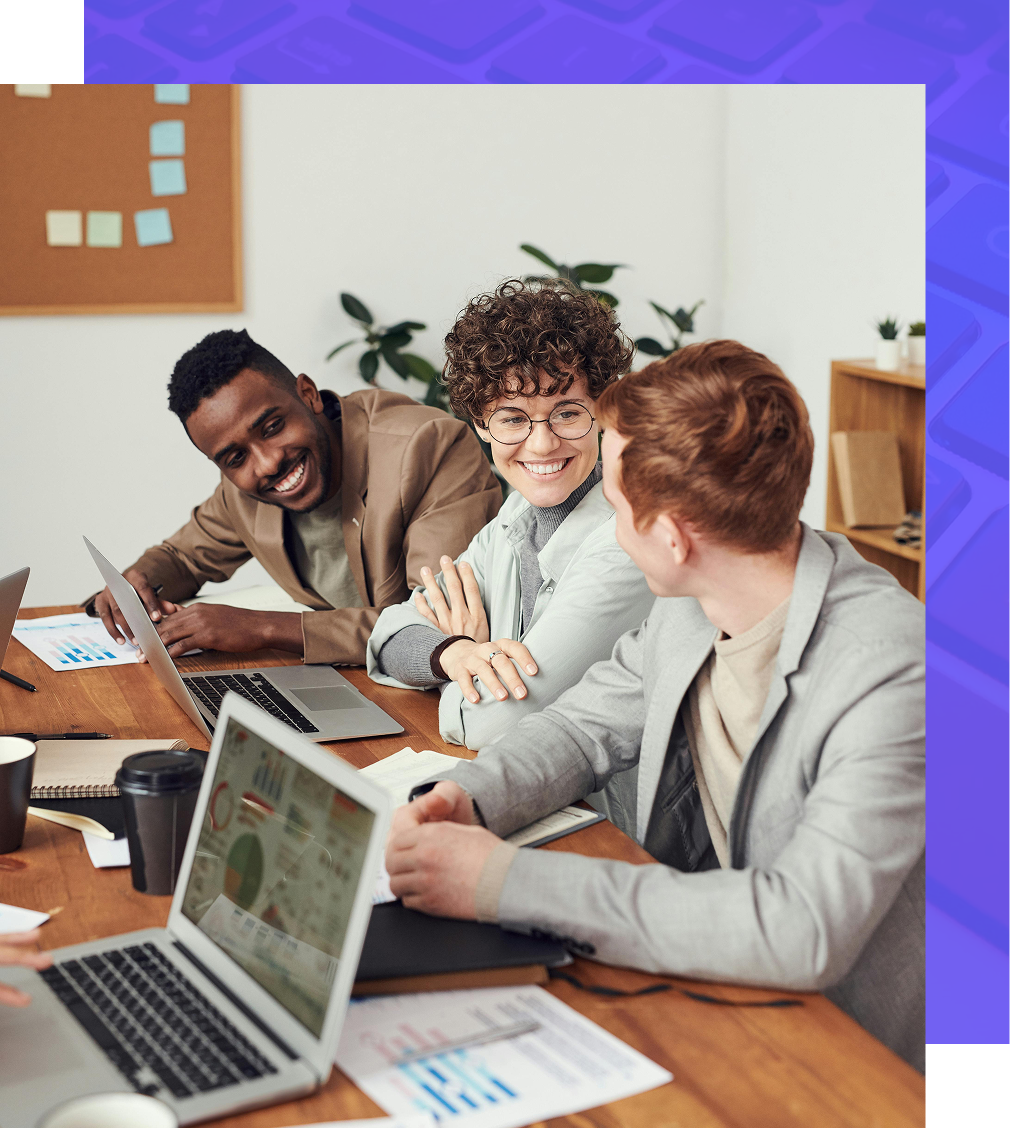 Three coworkers smiling and working together at a table with laptops and papers.
