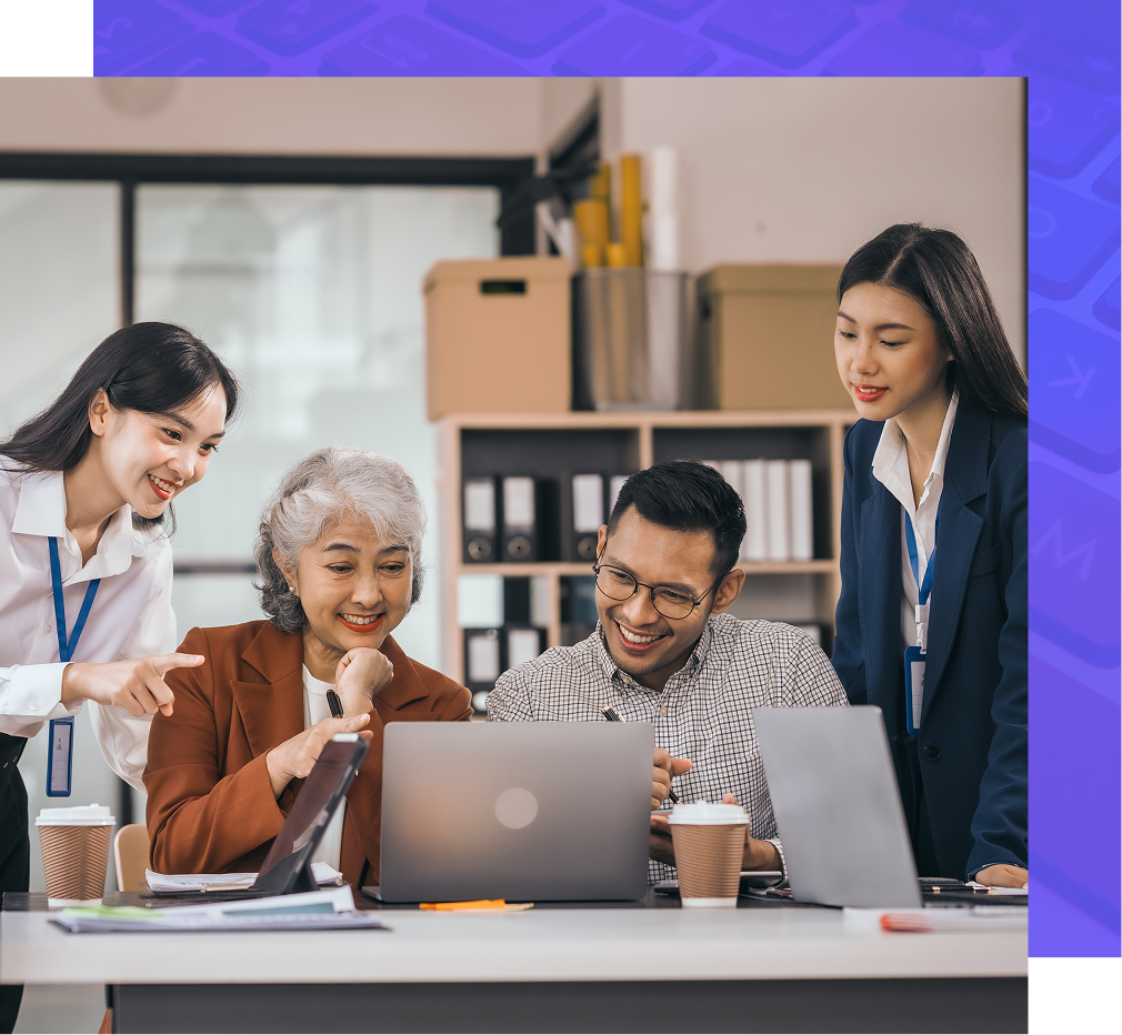 Four people collaborate around laptops in an office, smiling and pointing at the screens.