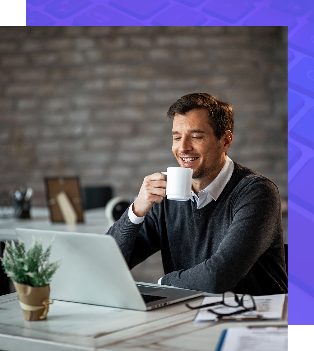 Man smiling, drinking from a mug, working on a laptop at a desk.