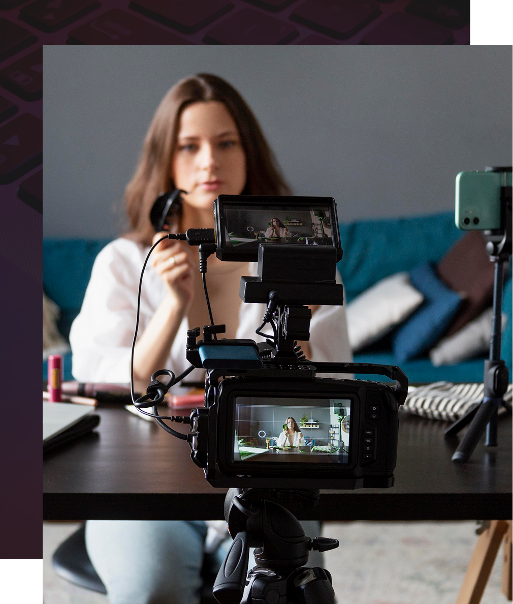 Woman filming a video with two cameras, applying makeup at a desk, teal couch in the background.