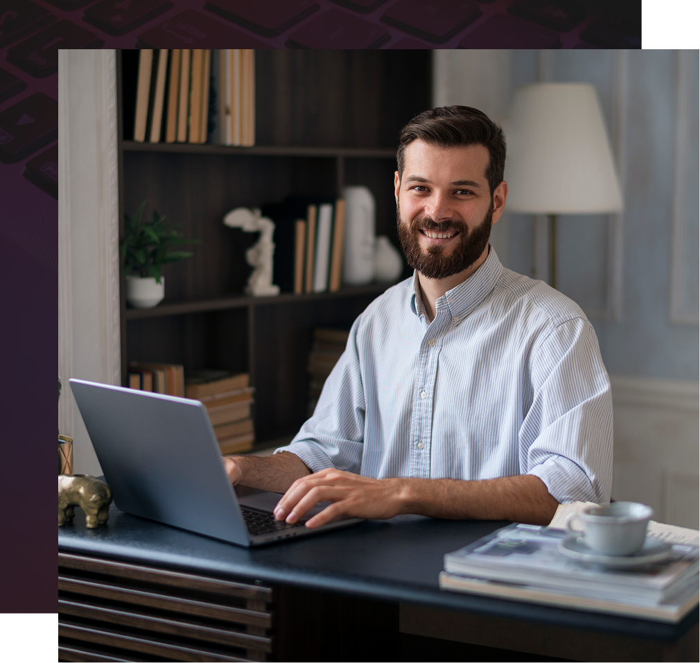 Man with beard smiling while working on laptop at a desk in home office, books in background.