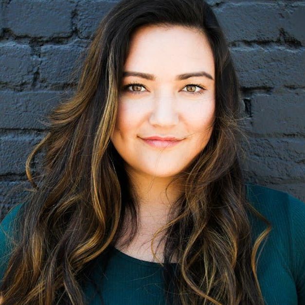 A woman with long hair is smiling in front of a brick wall.