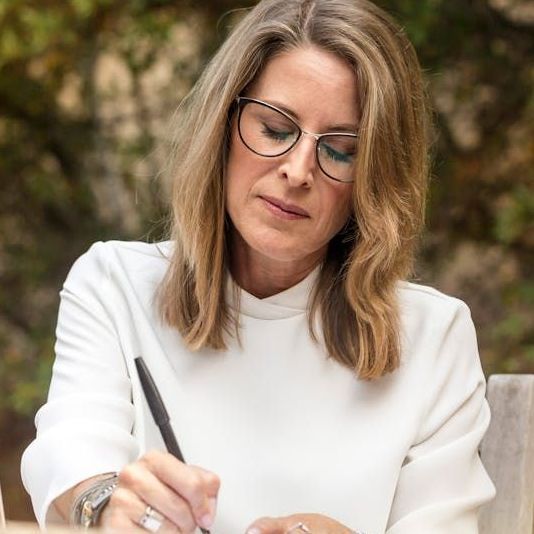 A woman wearing glasses and a white shirt is writing on a piece of paper.