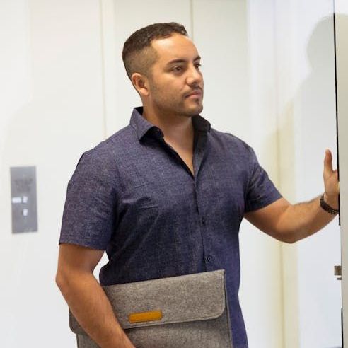 A man in a blue shirt is holding a briefcase and leaning against a wall.