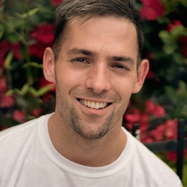 A man in a white shirt is smiling in front of red flowers