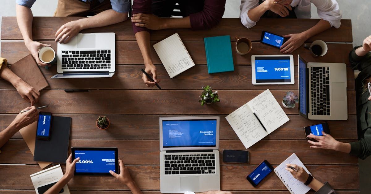 A group of people are sitting around a wooden table with laptops and tablets.