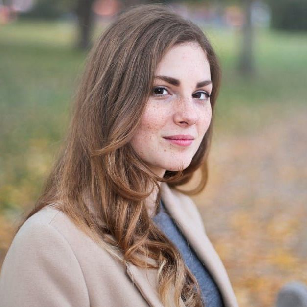 A woman in a tan coat is sitting in a park and looking at the camera.