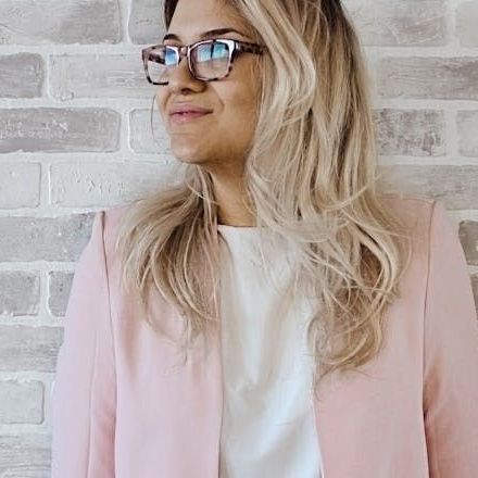 A woman wearing glasses and a pink jacket is standing in front of a brick wall.