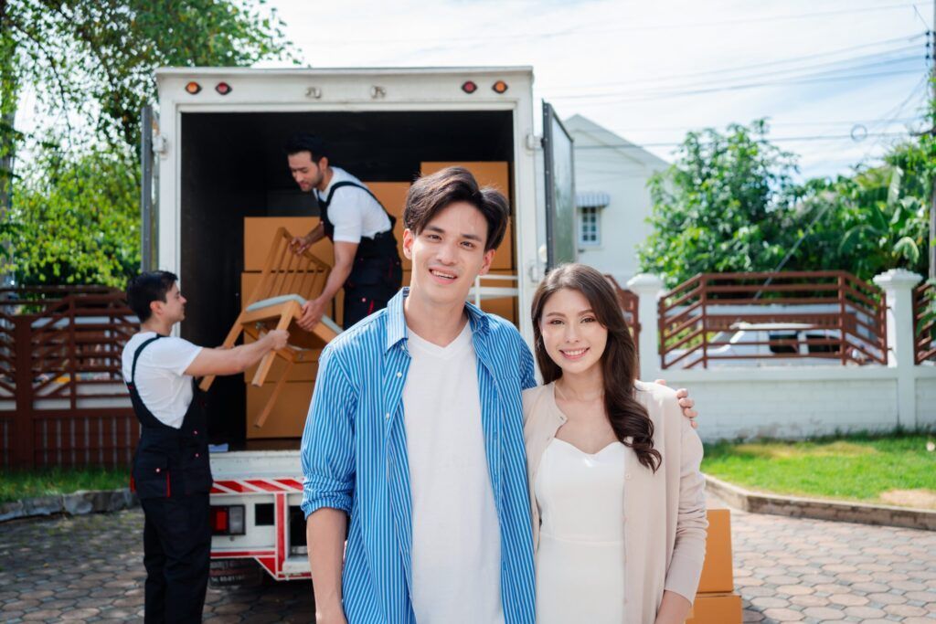 A smiling couple stands in front of a moving truck while workers unload cardboard boxes in a residential driveway.
