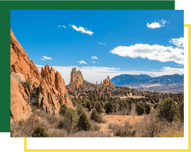 Red rock sandstone formations rise above a desert landscape with scrub brush under a blue sky with clouds.
