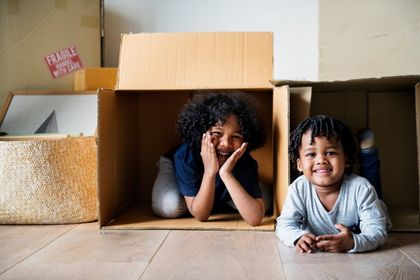 Two smiling children play inside cardboard boxes on a light wood floor in an indoor setting.