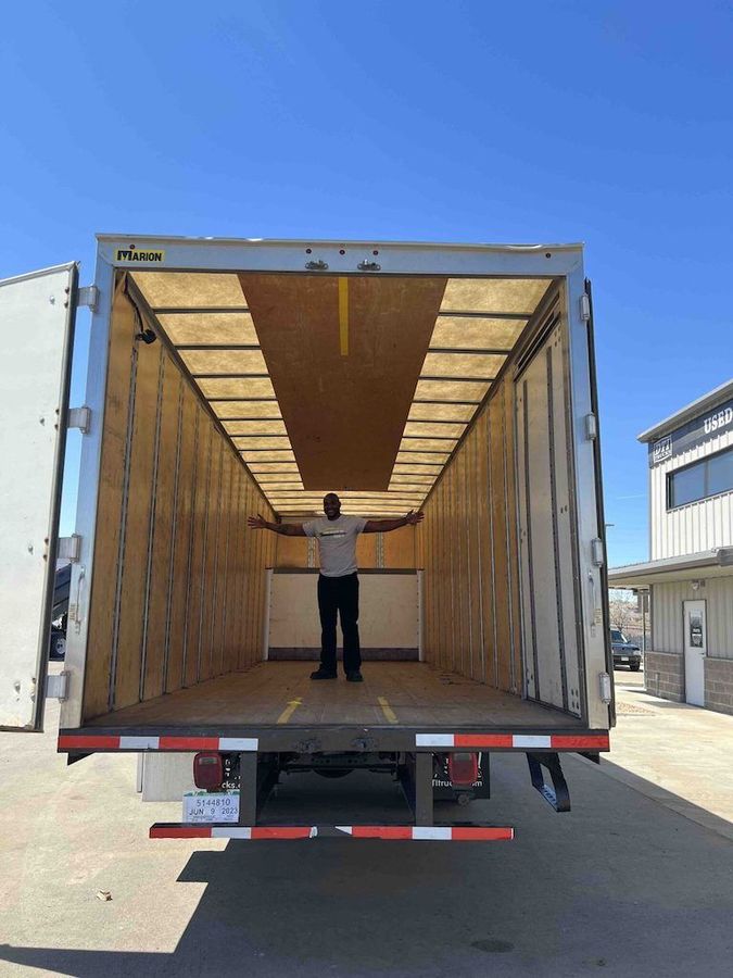A person stands with arms outstretched inside an empty, rectangular box truck trailer on a sunny day.