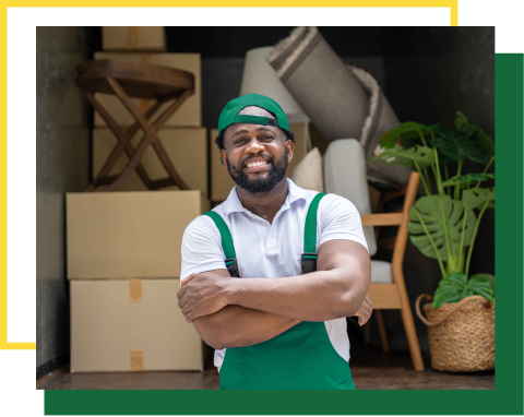 A smiling professional mover in a green uniform stands with arms crossed inside a truck filled with packed furniture.