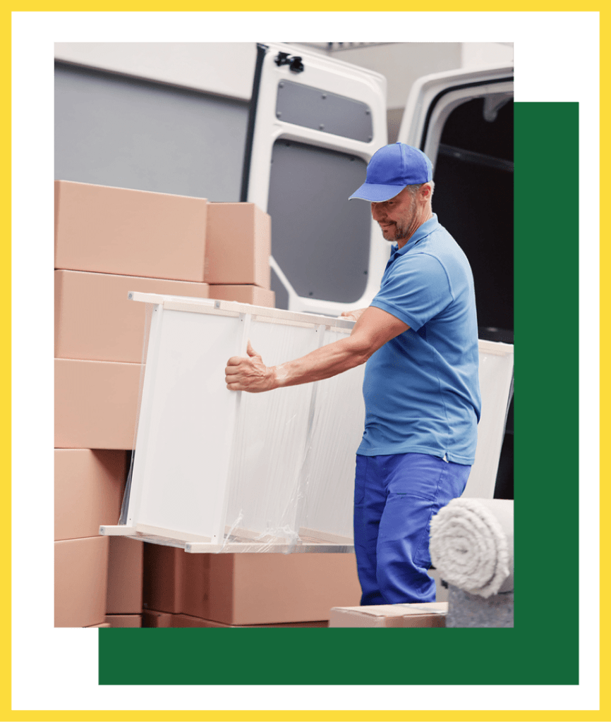 A person in a blue uniform carries a large white rectangular item out of the back of a van filled with cardboard boxes.