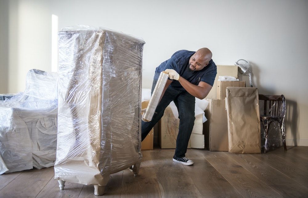 A person in a room preparing furniture for a move by wrapping a tall, wrapped item in protective plastic.