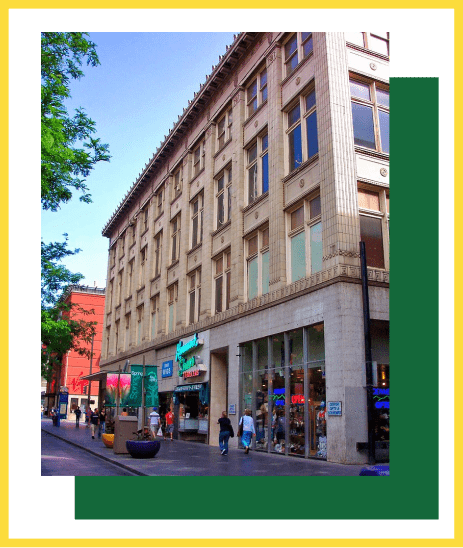 A tan, multi-story commercial building on a city street with pedestrians walking by storefronts under a clear blue sky.