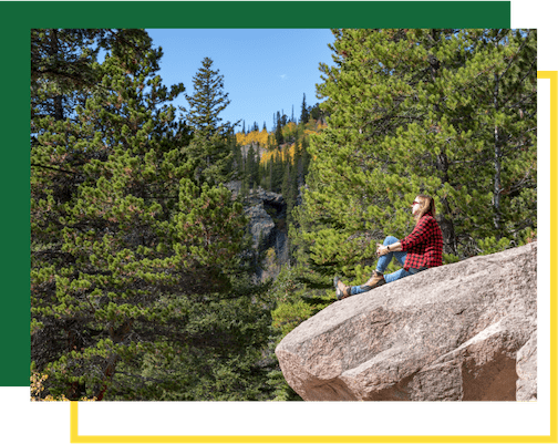 A person in a red plaid shirt sits on a large, tan boulder, looking out at a forest with tall evergreen trees.