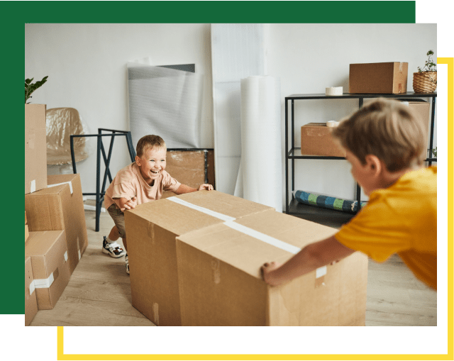 Two children pushing cardboard boxes across a floor in a room filled with packing supplies.