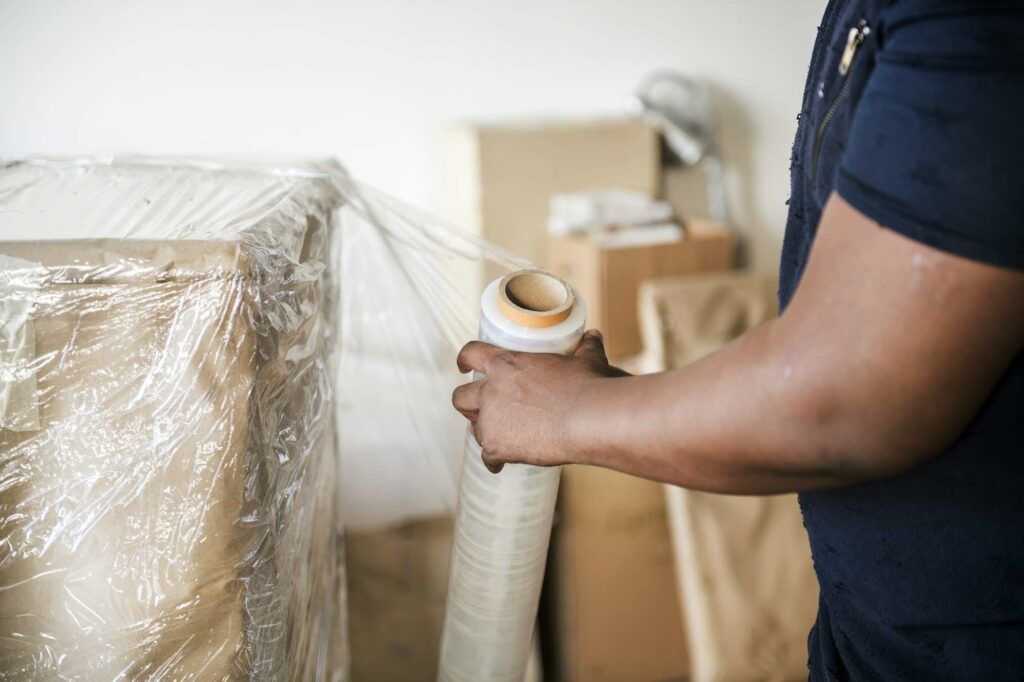 A person wraps a stack of cardboard boxes in plastic stretch film for storage or transport.