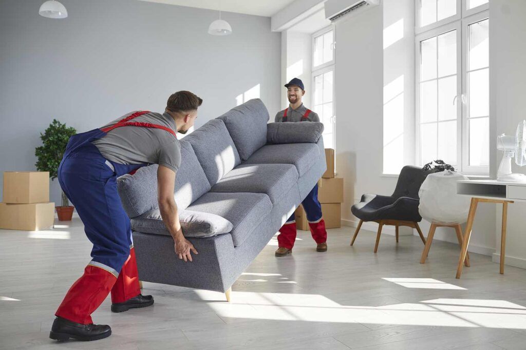 Two movers in blue and red uniforms carry a gray sofa across a bright, modern living room with large windows.