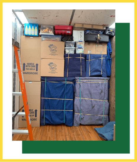 A moving truck interior packed with organized cardboard boxes, furniture wrapped in blue blankets, and a ladder to the left.