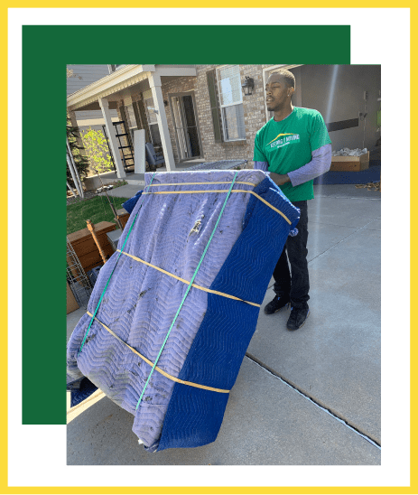 A mover in a green shirt carefully transports a large, blue-wrapped piece of furniture on a dolly outside a home.