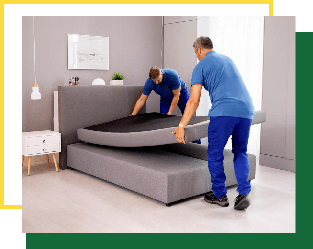 Two workers in blue uniforms lift the mattress of a grey upholstered bed frame in a modern bedroom.
