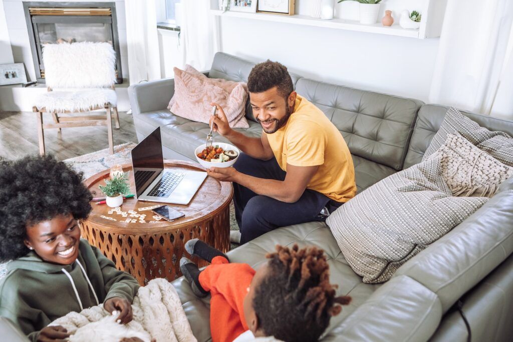 A family relaxes in a living room, with one person eating a meal on a sofa near a coffee table with a laptop.