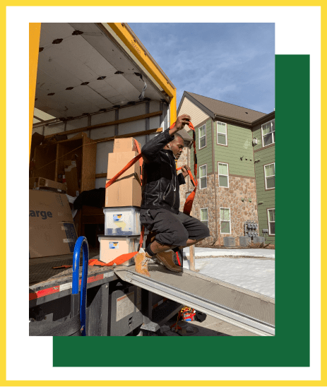 A person secures a stack of moving boxes inside a truck with a strap while kneeling on the vehicle's loading ramp.