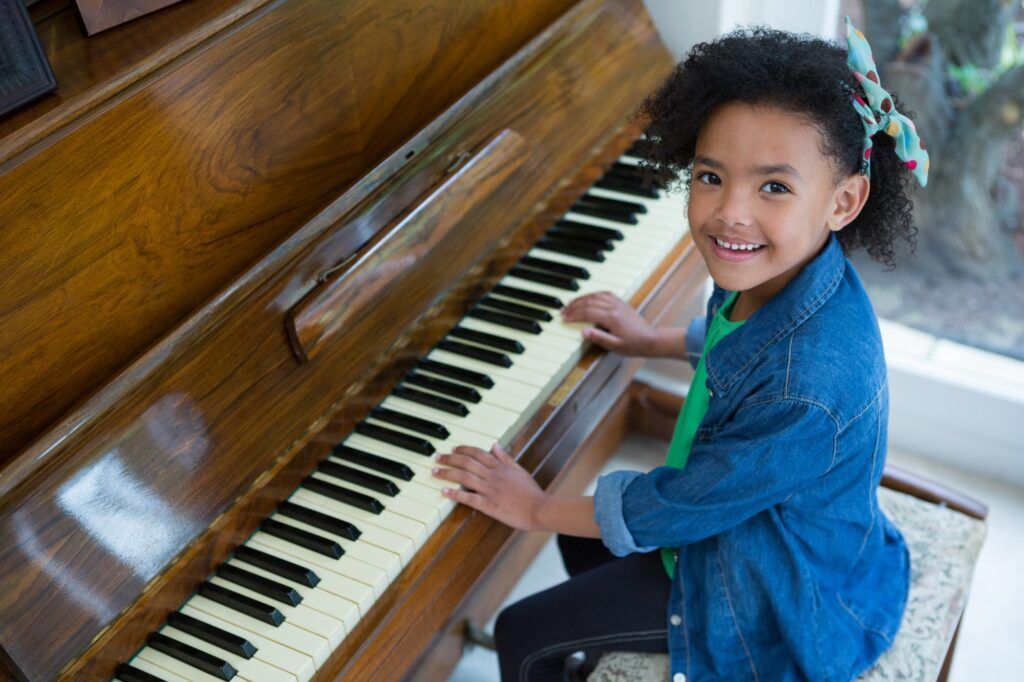 A smiling person with curly hair and a bow playing an upright wooden piano.